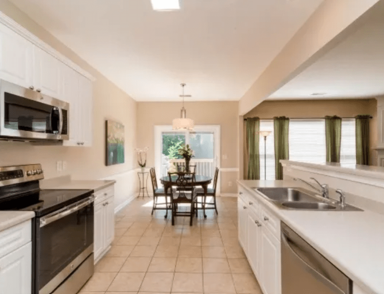 Bright kitchen featuring white cabinetry, stainless steel appliances, and a dining area with a table and chairs. Natural light flows in from windows and a door.
