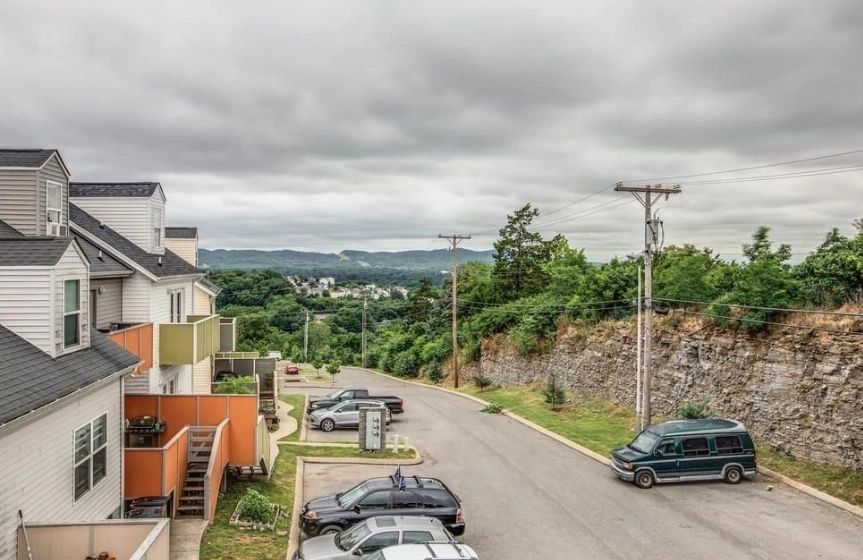 View of a residential street lined with parked cars, set against a backdrop of rolling hills under a cloudy sky.
