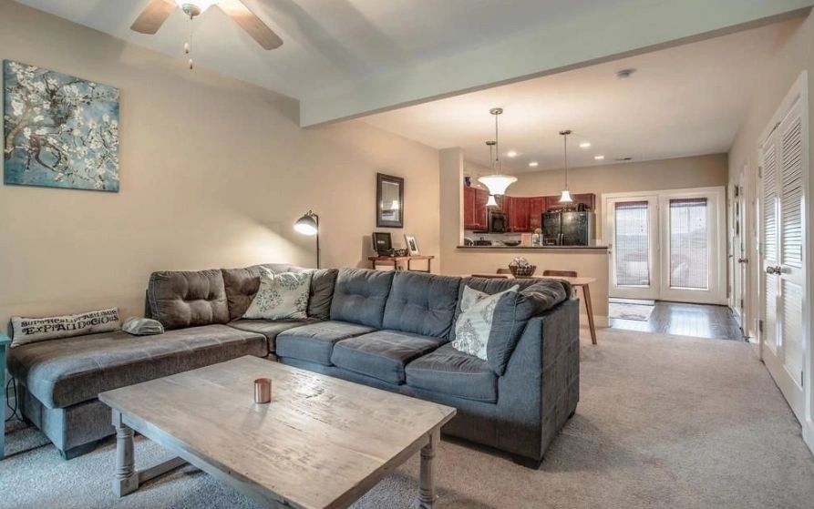 Cozy living room featuring a L-shaped gray sofa, a wooden coffee table, and an open kitchen view. Natural light floods in through patio doors.
