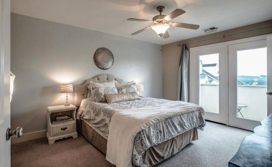 Cozy bedroom featuring a queen bed, stylish bedding, and a ceiling fan. Large windows open to a balcony with a scenic view.